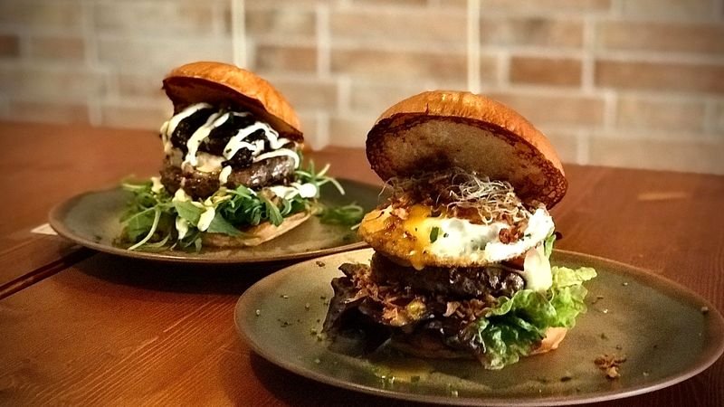 Dos hamburguesas de autor con pan brioche servidas en platos de piedra sobre una mesa de madera, con la pared de ladrillo del restaurante al fondo.