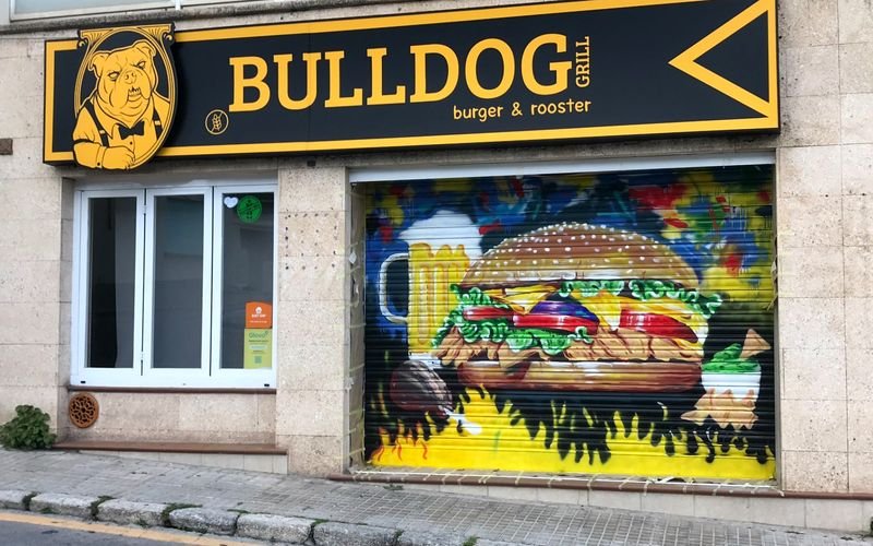 Fachada del restaurante con el rótulo amarillo de BULLDOG grill, burger & rooster, junto al mural pintado de la persiana.